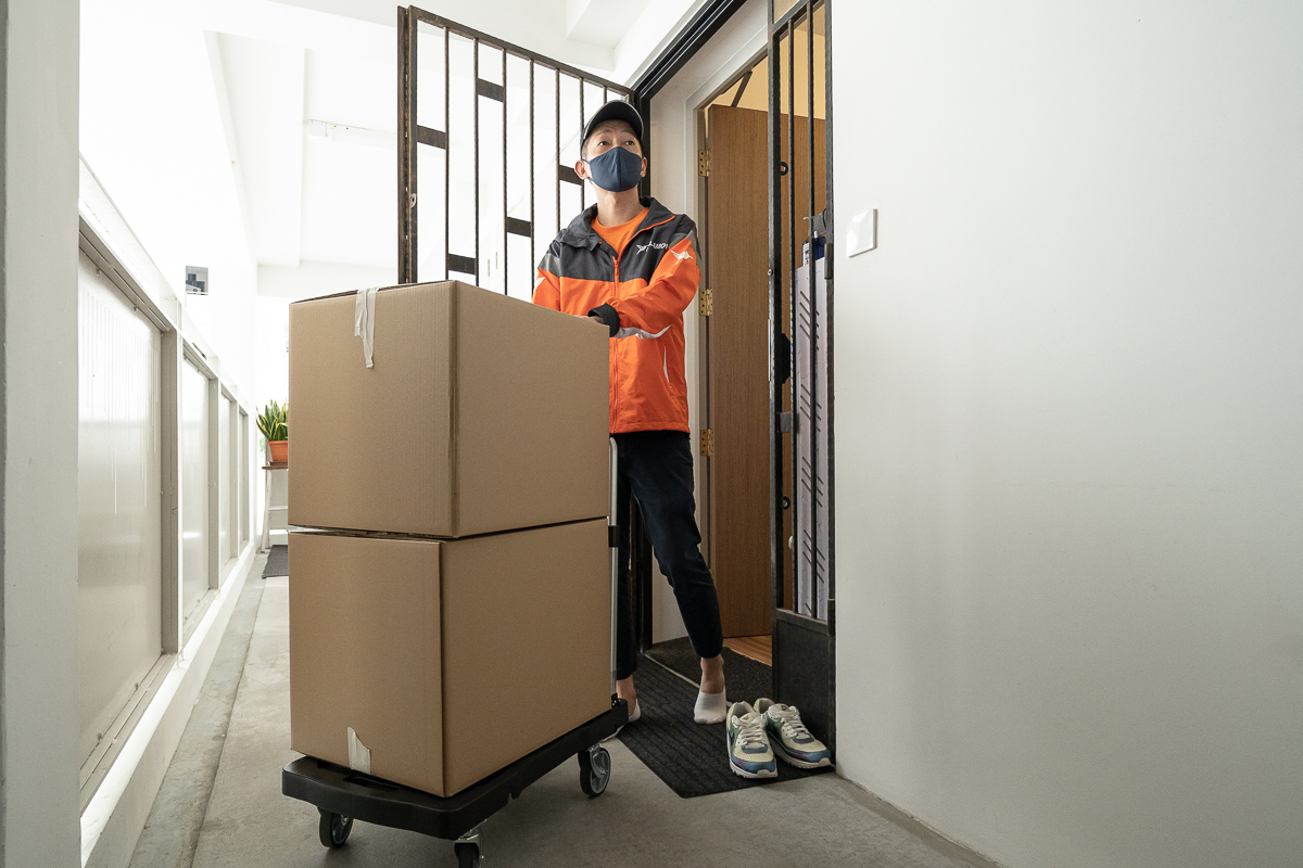 squarerooms lalamove moving service furniture belongings delivery boxes new home transport singapore bernard lim model man in orange uniform standing in apartment hdb gate with boxes on trolley and crossed arms